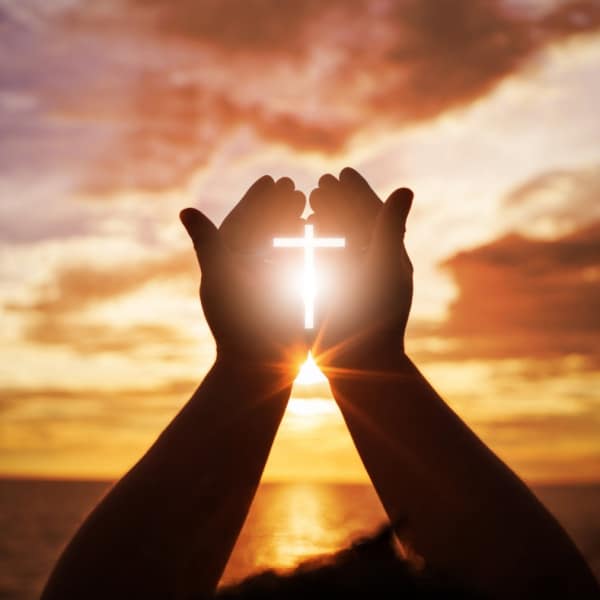 Hands reaching towards sun while holding a cross